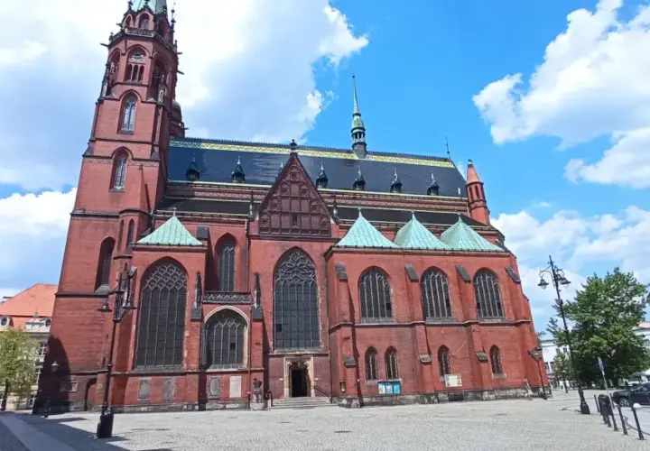 Gotycka katedra w mieście nad Kaczawą, z ceglaną fasadą i zielonymi dachami, na tle błękitnego nieba z chmurami.