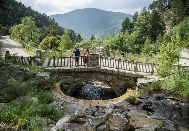 Pareja en un puente de piedra sobre un arroyo en Les Pardines, Andorra. Montañas y bosques al fondo.