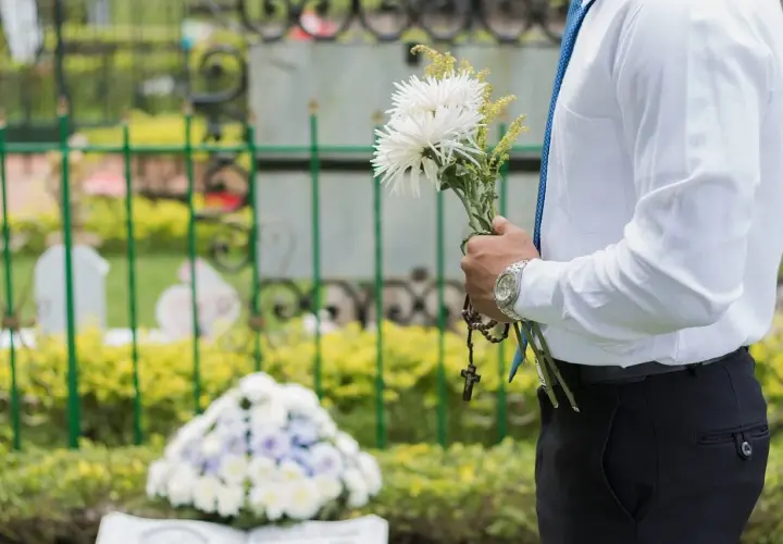 Mężczyzna w białej koszuli i niebieskim krawacie trzyma bukiet białych kwiatów i różaniec. W tle cmentarz, gdzie spoczywaj w pokoju.