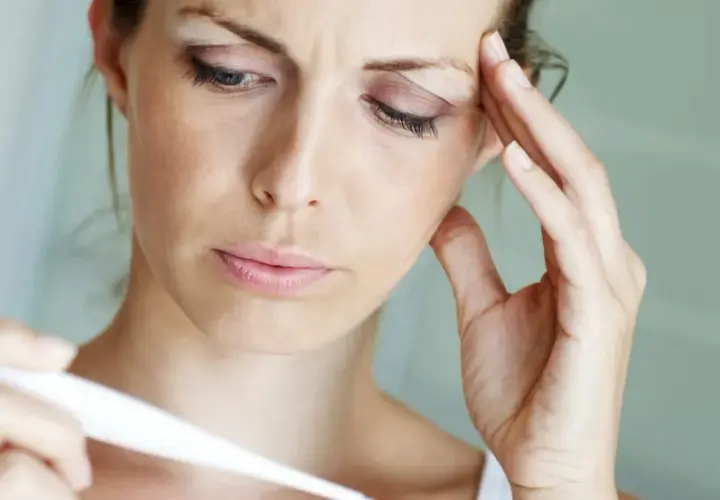 Frau mit Fieber, die sich die Stirn hält und ein Thermometer betrachtet. Psychosomatischer Fieber kann viele Ursachen haben.