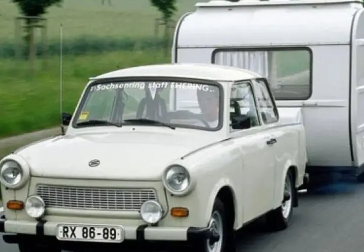 Biały trabant ciągnie przyczepę kempingową po drodze.