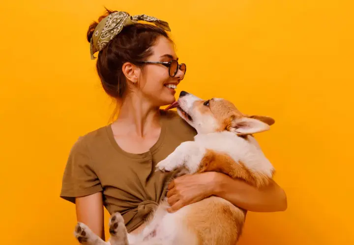 Uśmiechnięta dziewczyna z okularami i bandaną na głowie tuli corgi. Piesek liże ją po nosie. Wiele imion na N, jak Nela czy Nika, pasowałoby do tej słodkiej pary.