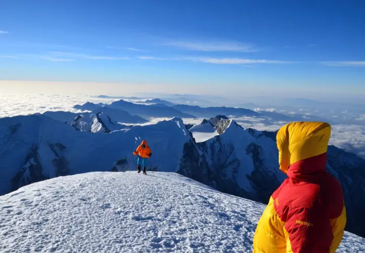 Widok z góry, gdzie są Himalaje. Dwóch alpinistów w kolorowych kurtkach na ośnieżonym szczycie, pod nimi morze chmur i kolejne pasma górskie.