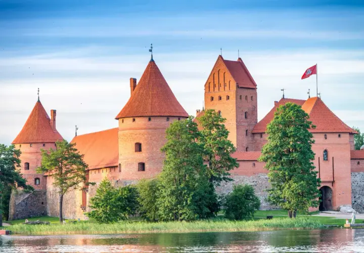 Zamek w Trokach, majestatyczna ceglana twierdza z czerwonymi dachami, wznosi się nad wodą, otoczony zielenią.