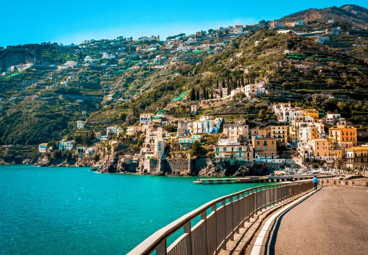 A scenic road curves along the coast, offering breathtaking views of colorful buildings clinging to a hillside. The turquoise sea sparkles below, a perfect backdrop for an unforgettable Amalfi drive.