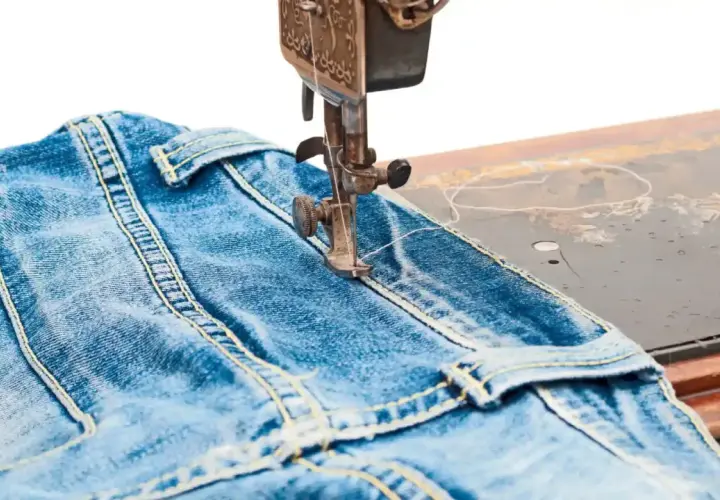 A sewing machine stitches a classic jean stitch onto blue denim fabric, highlighting the durable construction of jeans.