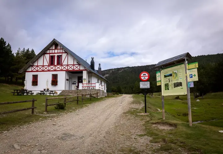 Refugi comes de rubio, un refugio de montaña blanco con detalles rojos, rodeado de pinos y un camino de grava.