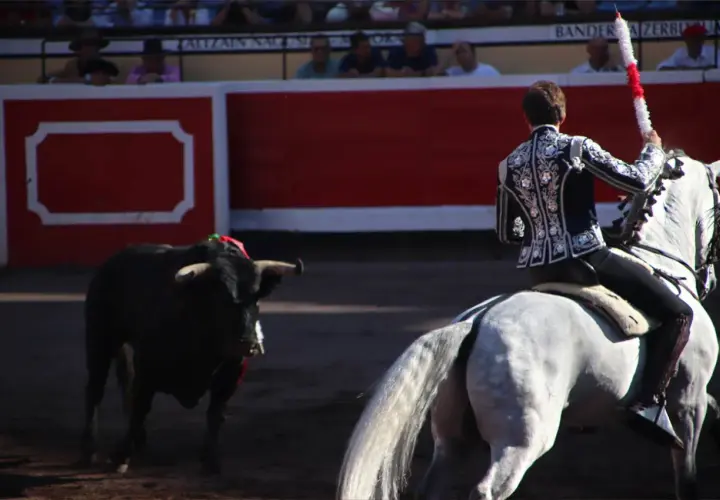 Un torero a caballo se prepara para el lance. El toro, con adornos en la cabeza, espera en la arena. El tendido alto las ventas, lleno de público, observa expectante.