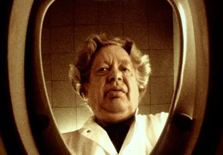 En man med lockigt hår, klädd i vit rock, tittar in i en toalettstol. Ernst Hugo Järegård i en minnesvärd scen.