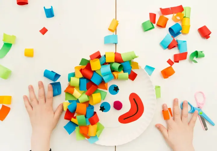 Bambini creano lavoretti di carnevale da appendere: un pagliaccio con capelli colorati su un piatto di carta, circondato da coriandoli di carta.
