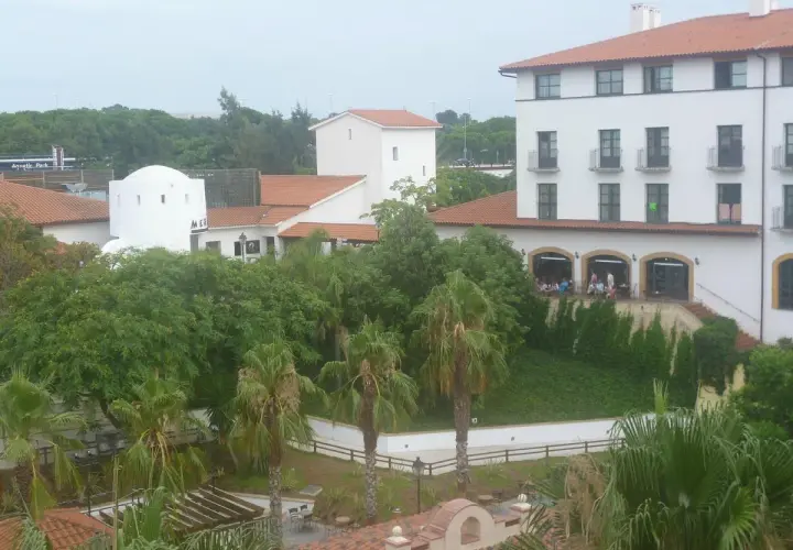 Edificio blanco con tejado naranja, palmeras y vegetación exuberante. Parece el hotel El Paso PortAventura, listo para recibir a sus huéspedes.