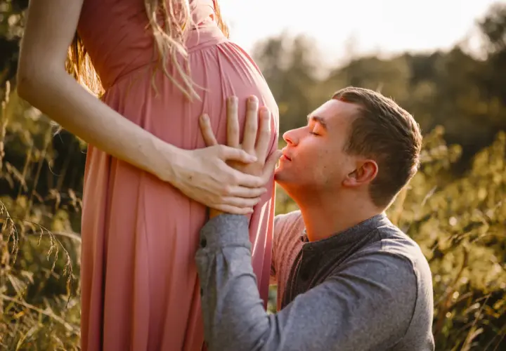 Mężczyzna całuje brzuch ciężarnej kobiety w różowej sukience na tle natury.
