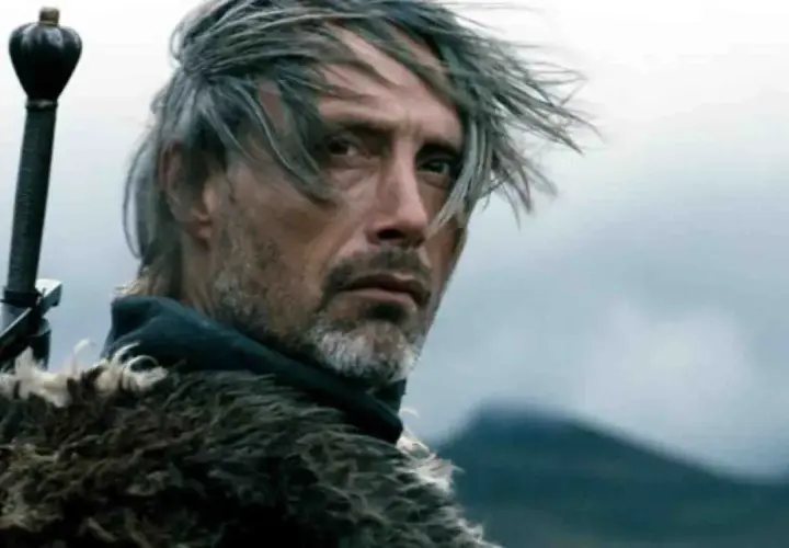 Mads Mikkelsen młody, z mieczem za plecami i w futrzanym płaszczu, spogląda w dal.