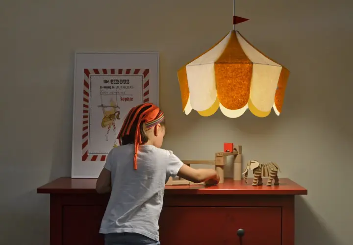 Una niña juega con bloques de madera bajo una lámpara colgante infantil con forma de carpa de circo.