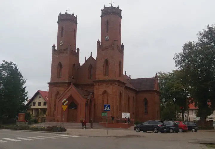Imponujący kościół w Krokowej z cegły, z dwiema wieżami i zegarami, otoczony zielenią i samochodami.
