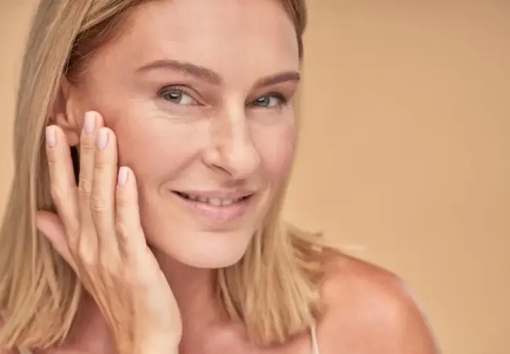 Uśmiechnięta kobieta z blond włosami dotyka policzka, prezentując naturalny wygląd skóry dzięki dobremu podkładowi kryjącemu bez efektu maski.