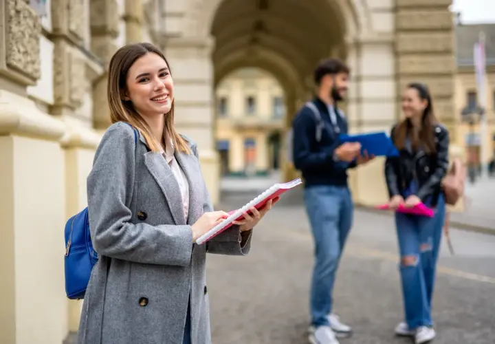 Uśmiechnięta studentka z notatnikiem w ręku, przygotowująca się do studiów 2 stopnia. W tle para studentów.