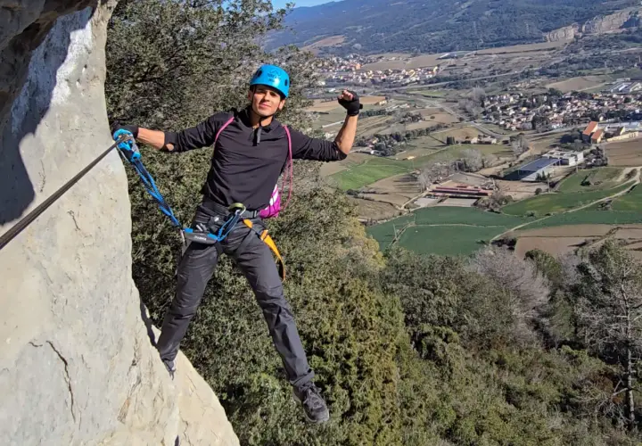 Un escalador sonriente, con casco azul, disfruta de la vía ferrata, un ejemplo de los diversos tipos de escalada, con un pueblo y campos verdes de fondo.