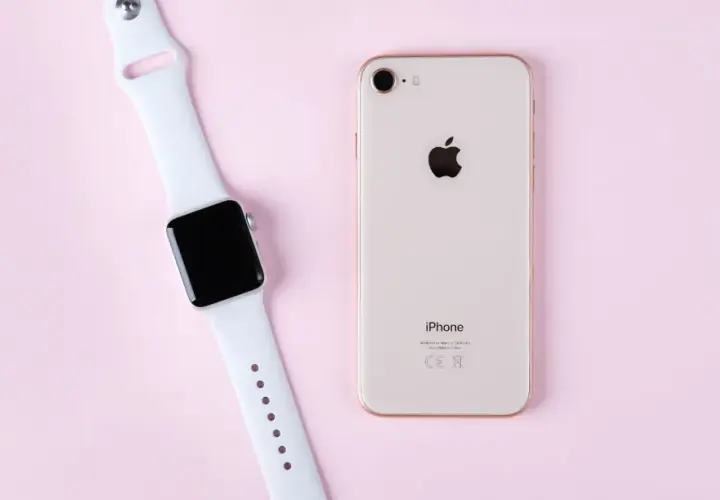 Apple Watch und iPhone liegen nebeneinander auf rosa Untergrund, bereit zum Laden.