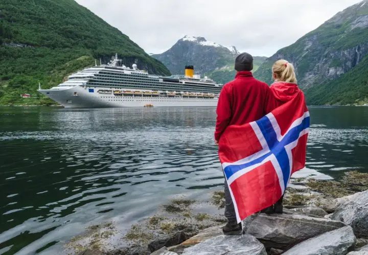 Para podróżuje po Norwegii, z flagą narodową i statkiem wycieczkowym w tle. Planujesz podróż? Pomyśl o ubezpieczenie Norwegia.