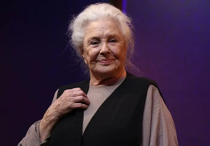 Lola Herrera, con su melena plateada y una sonrisa serena, luce un chaleco negro sobre una blusa beige. Su mano izquierda, con un anillo, reposa sobre su pecho.