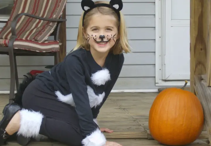 Bambina con trucco da gatto e orecchie, pronta per vestiti carnevale veloci fai da te bambini. Zucca arancione accanto.