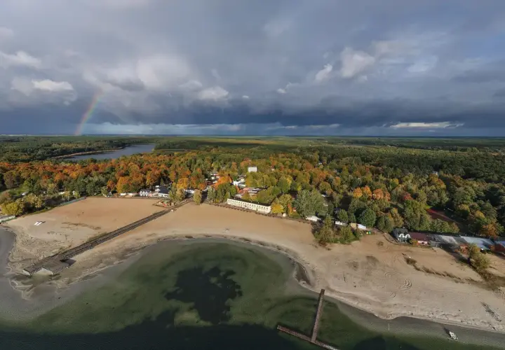 Jesioro Skorzęcin jesienią. Tęczowy łuk nad ciemnymi chmurami, piaszczysta plaża i kolorowy las.