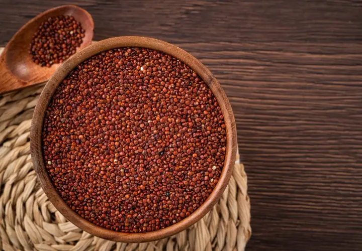 Una ciotola di legno piena di quinoa rossa, con un cucchiaio di legno accanto, su un sottobicchiere intrecciato.