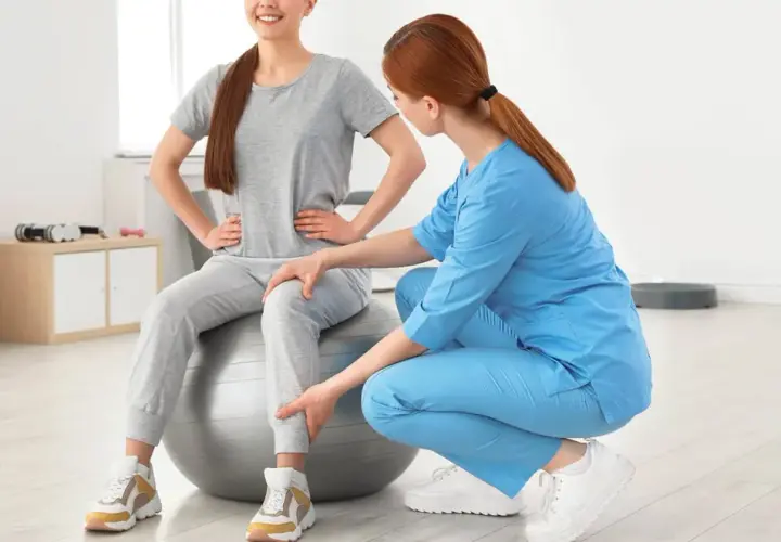 Une kinésithérapeute aide une patiente assise sur un ballon de gym à retrouver de la force dans les jambes grâce à des exercices ciblés.