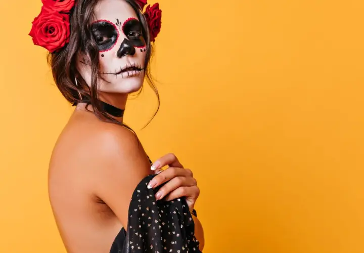 Kobieta z makijażem sugar skull i czerwonymi różami we włosach na pomarańczowym tle.