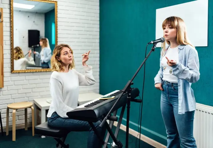 Lekcja śpiewu w domu: nauczycielka przy keyboardzie instruuje uczennicę przed mikrofonem.