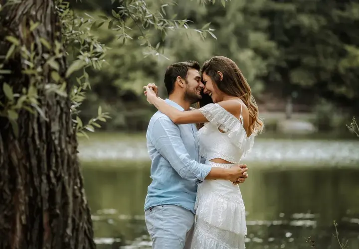 Para młoda, w białej sukni i błękitnej koszuli, przytula się czule nad jeziorem. Ich uśmiechy i spojrzenia sugerują szczęśliwą noc poślubną.
