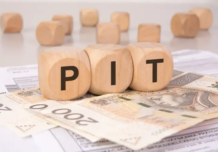 Drewniane klocki z literami "PIT" leżą na banknotach i dokumentach. Może to symbolizować rozliczenie podatkowe lub umowę darowizny.
