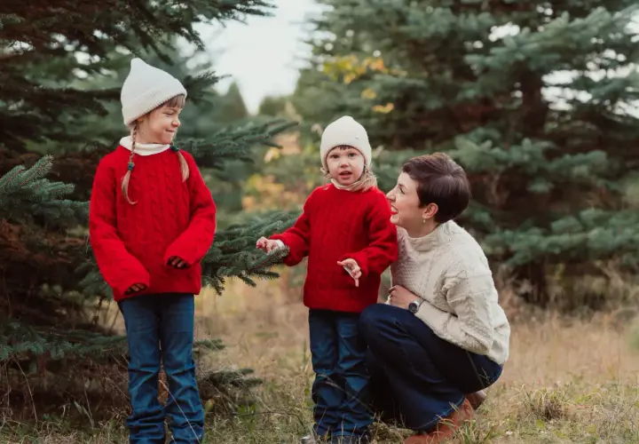 Urocza sesja zdjęciowa dla dziecka w otoczeniu choinek. Mama i dwie córki w czerwonych sweterkach i białych czapkach cieszą się wspólnym czasem.