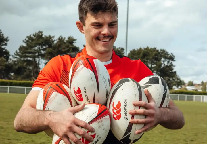 Uśmiechnięty mężczyzna w czerwonej koszulce trzyma kilka piłek do rugby i futbolu amerykańskiego.