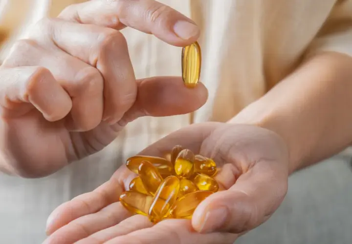 Dłoń trzyma garść kapsułek z olejem rybim, bogatym w kwasy omega 3. Jedna kapsułka jest podniesiona między palcami.