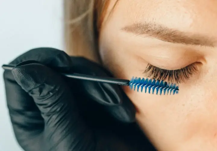 Stylist w czarnej rękawiczce szczotkuje przedłużone rzęsy. Zastanawiasz się, jak długo odrastają rzęsy po przedłużaniu?