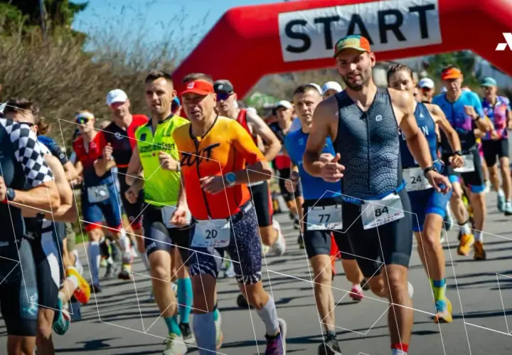 Zawodnicy biegną pod czerwoną bramką startową. Wszyscy przestrzegają zasad triathlonu, skupieni na rywalizacji.