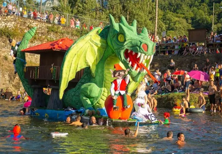 Dragón verde gigante en un desfile acuático, parte de las fiestas de prao Asturias. Gente en el agua y espectadores en la orilla.
