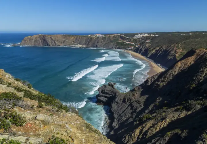 Malownicze wybrzeże Portugalii z klifami, piaszczystą plażą i turkusowym oceanem. Białe fale rozbijają się o brzeg, a w oddali widać białe domki.