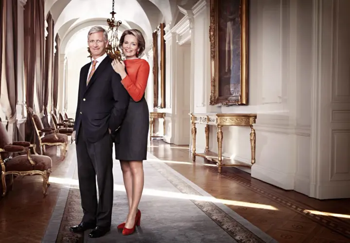 Królowa Belgii, uśmiechnięta, stoi obok króla w eleganckim korytarzu pałacowym.