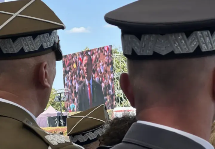 Dwóch mężczyzn w mundurach, jeden z nich ma jedną gwiazdkę na pagonie, ogląda przemówienie na telebimie.