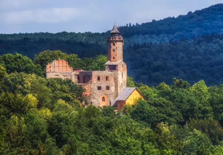 Ruiny zamku na wzgórzu otoczone zielenią, w oddali widać las i niebo. Widok przypomina krajobraz nad jeziorem Bystrzyckim.