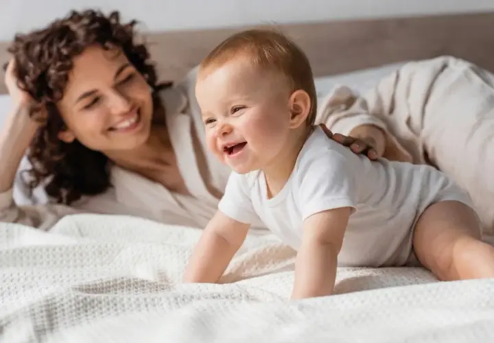 Un feliz bebe 7 meses gatea en la cama mientras su madre sonríe.