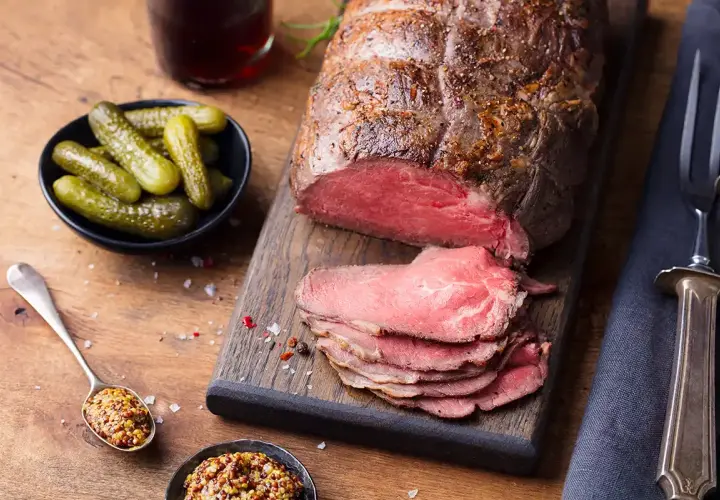 Un rôti de bœuf tranché, servi avec des cornichons, de la moutarde et des couverts sur une planche en bois.
