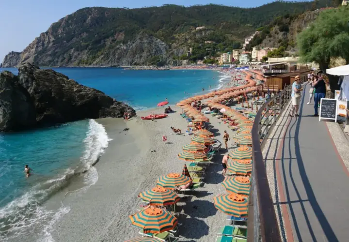 Plage animée à Moneglia, Italie, avec des parasols rayés, des baigneurs dans la mer turquoise et des collines verdoyantes en arrière-plan.