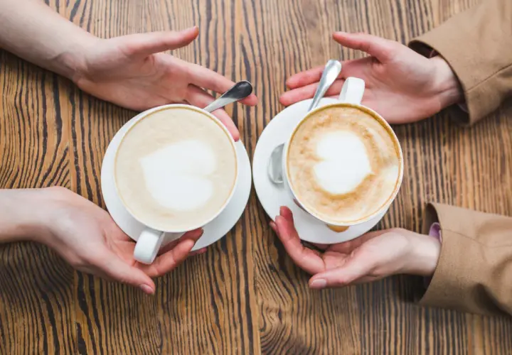 Zwei Cappuccinos mit Herz-Latte-Art, die Hände reichen sie sich über einen Holztisch. Wie viel Milch ist im perfekten Cappuccino?