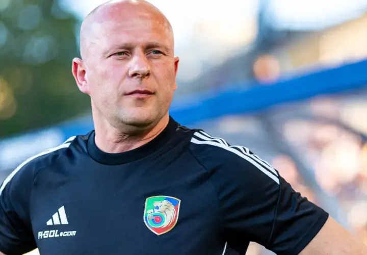 Łysy mężczyzna w czarnej koszulce z herbem Miedzi Legnica. Trener Miedzi Legnica w skupieniu obserwuje boisko.