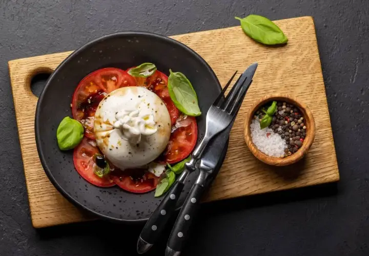 Pyszny **burrata przepis** na talerzu z pomidorami i bazylią. Obok miseczka z solą i pieprzem.