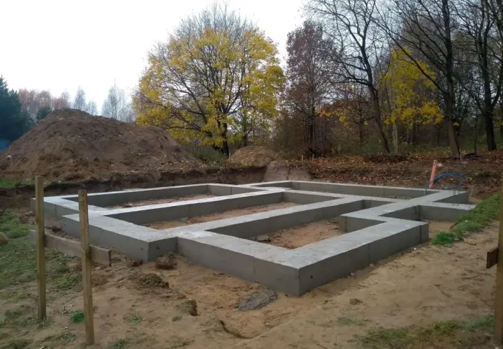 Fundament domu wylewany na ławach fundamentowych, poniżej strefa przemarzania gruntu. W tle jesienne drzewa i usypana ziemia.